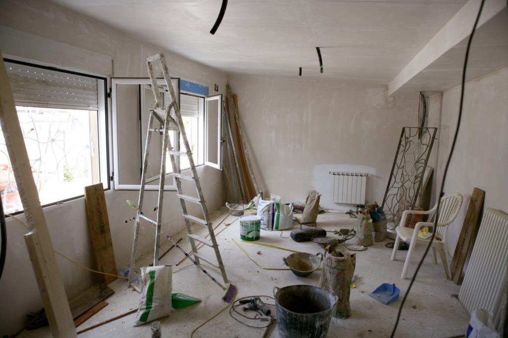 Image shows room in a house being remodeled. There is electrical wires hanging from the ceiling awaiting fixtures, a ladder, garbage can, dust pan, plastic chair, broom, cement bags, a wall heater and dust is visible. The windows are opened to allow ventilation.