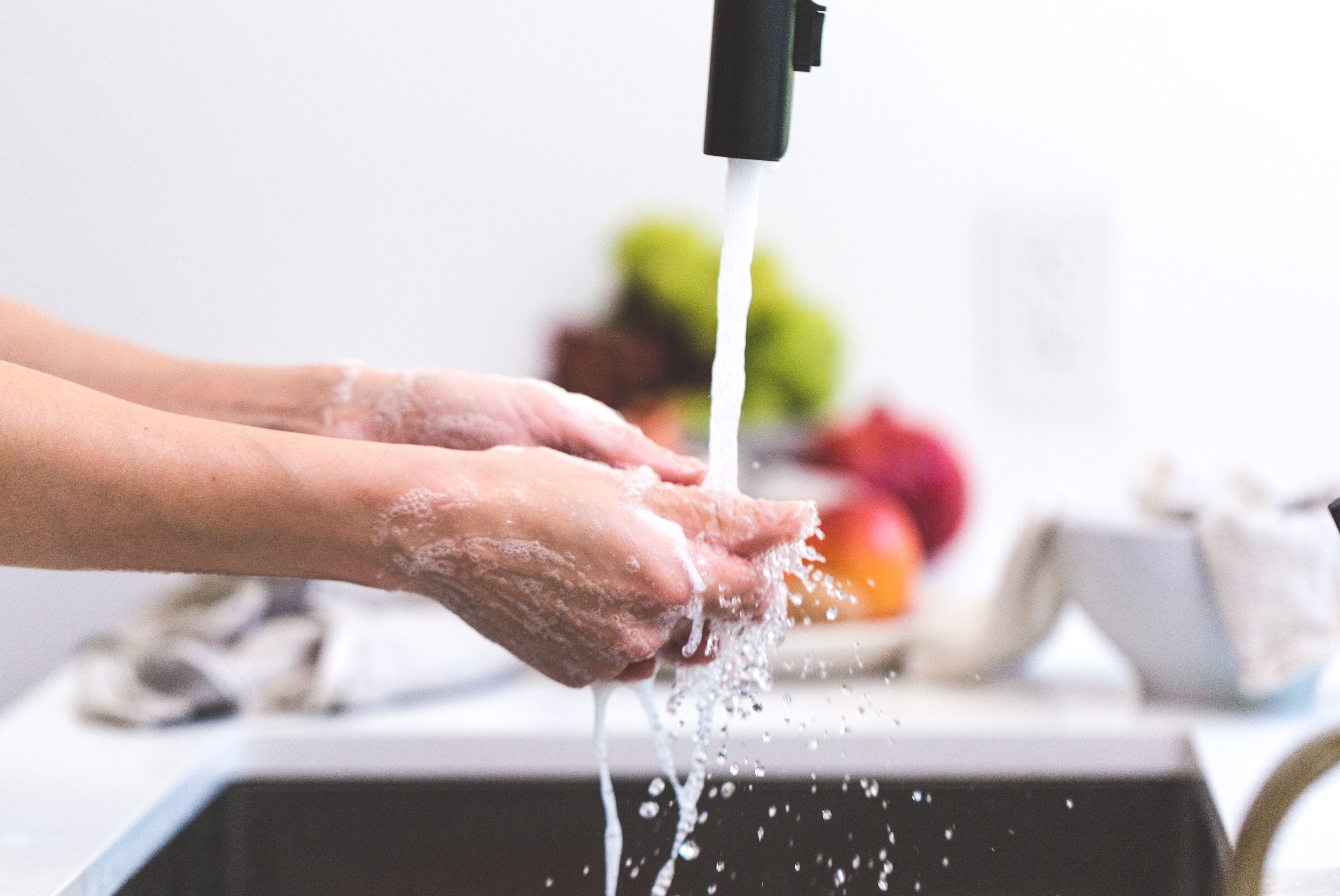 Image depicts a pair of hands being washed underneath a kitchen faucet with a bowl of fruit in the background