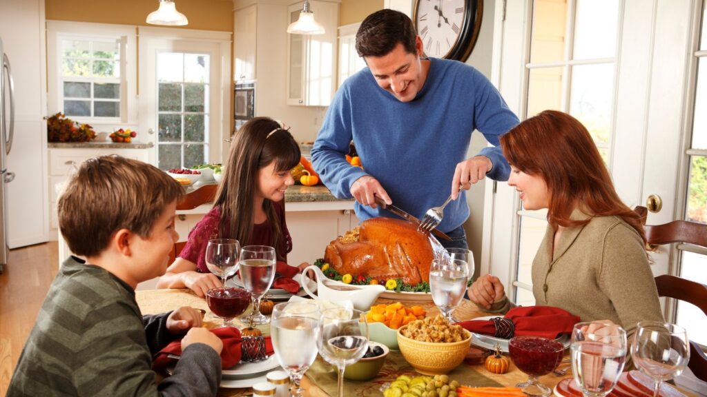 a family gathered around the Thanksgiving table while dad cuts a turkey.